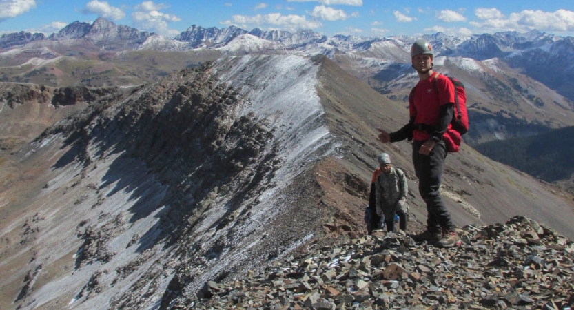 A person wearing safety gear stands atop a rocky summit in front of snow-capped mountains. Behind them, more people are making their way up to the summit. 